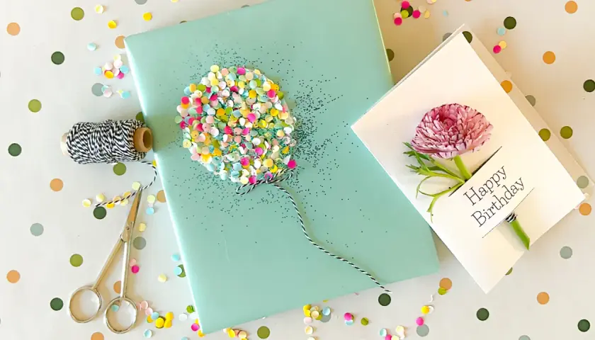 Ein liebevoll gestaltetes Geschenk zum 1. Geburtstag: Geschenkpapier mit Glitzer-Ballon, Schnur, Schere und eine Karte mit Blüte.