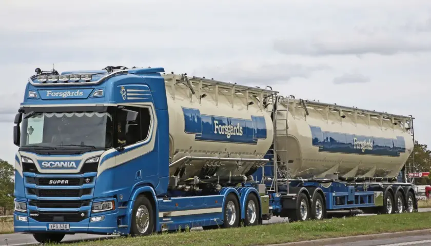 Ein blauer Scania-Sattelzug mit zwei Siloanhängern, beladen für Forsgårds, fährt auf einer schwedischen Straße. Das Tempolimit in Schweden ist zu beachten.