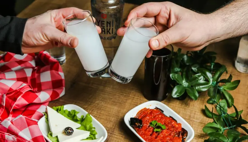 Dwie szklanki z białym alkoholem, raki, unoszą się do toastu. Na stole ser, sałatka i butelka Yeni Raki.