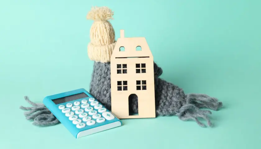 Casa de madera abrigada con bufanda y gorro, junto a calculadora. Simboliza el cálculo del coeficiente de rendimiento energético para el hogar.