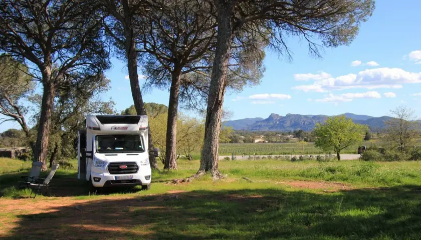 Un camping-car blanc est garé sous des pins, avec des chaises pliantes à côté. Un paysage de vignobles et de montagnes s'étend au loin. Un guide aire camping car idéal pour l'aventure.