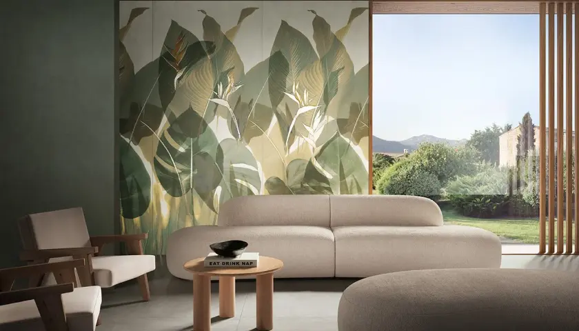 Soggiorno con divano beige e poltrone in legno, parete con fap rivestimenti a tema foglie tropicali e vista su giardino lussureggiante.