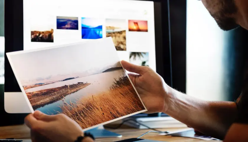 Fotograf przegląda wydrukowane zdjęcie krajobrazu, szukając inspiracji do nowych zleceń. Na ekranie komputera widać galerię jego prac.