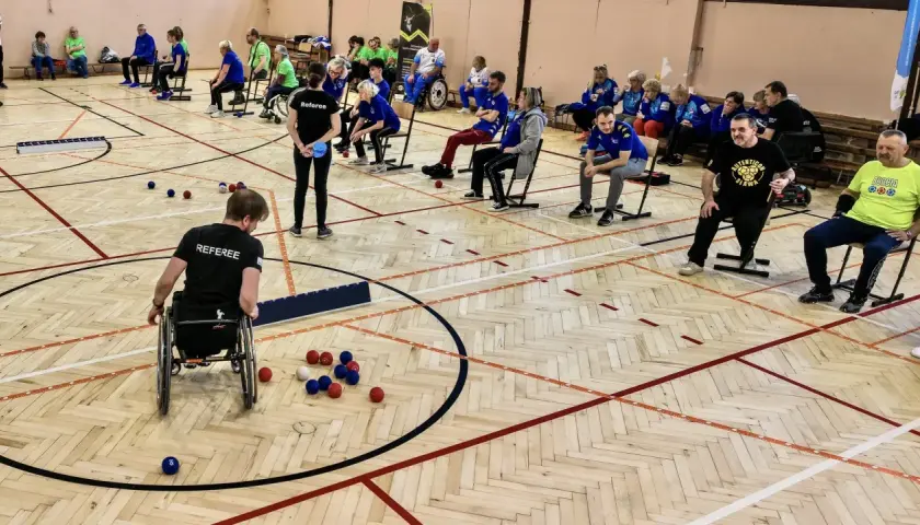 Zawodnicy grają w boccię, sportową grę precyzyjną. Na boisku widać kolorowe kule i zawodnika na wózku inwalidzkim.
