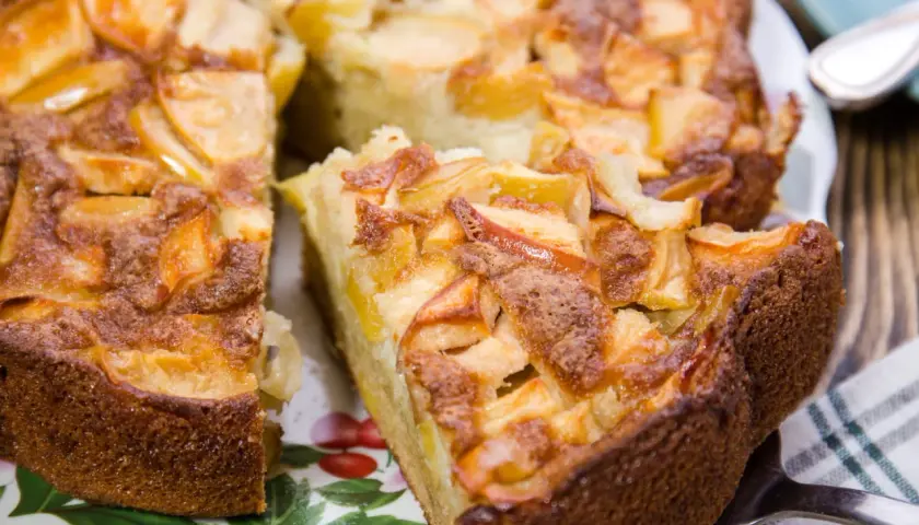 Un délicieux gâteau aux pommes, doré et moelleux, avec des morceaux de fruits caramélisés sur le dessus. Une part est déjà coupée, prête à être dégustée.