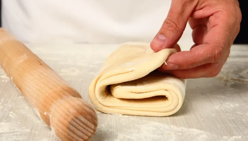 Mani che piegano pasta sfoglia, pronta per diventare pasta fatta in casa. Un mattarello è accanto, pronto per il prossimo passaggio.