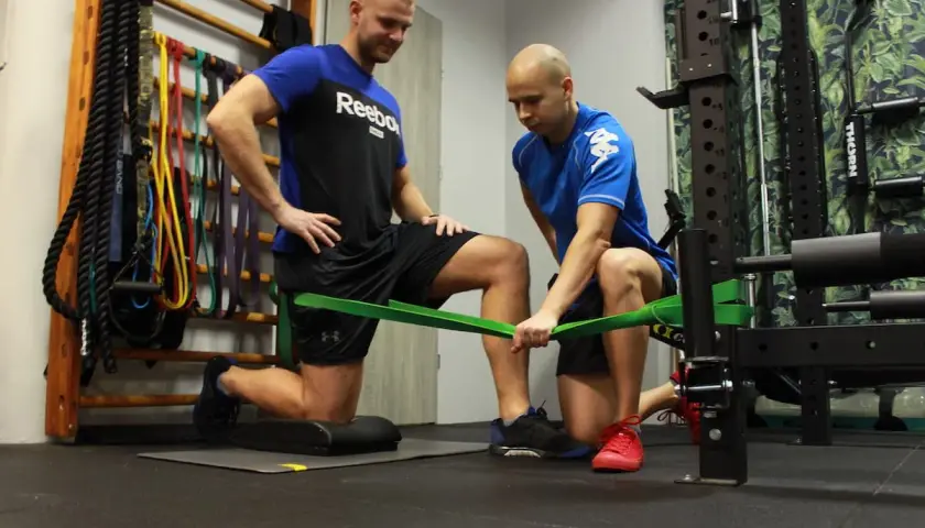 Trening dla par: jeden mężczyzna klęczy, drugi pomaga mu z taśmą oporową. W tle drabinki gimnastyczne i sprzęt do ćwiczeń.