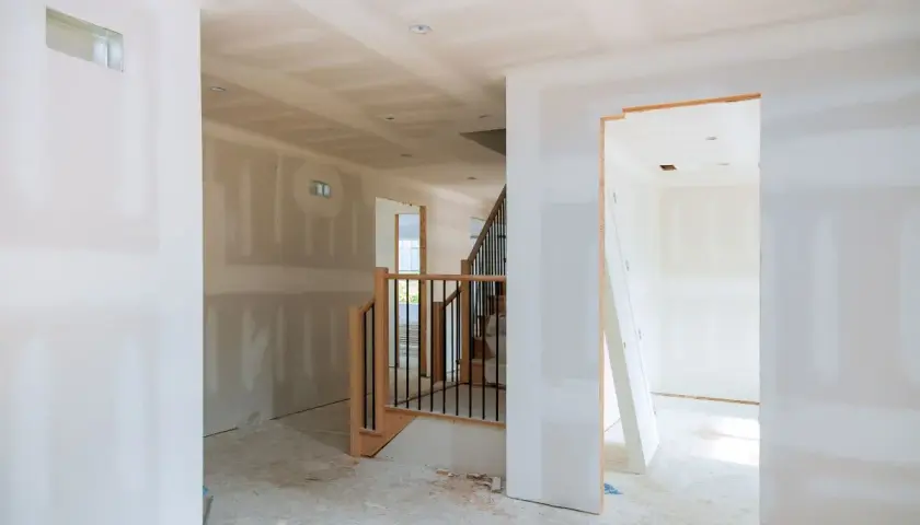 Interno di una casa in costruzione con pareti in cartongesso grezzo, una scala in legno e metallo e un'apertura verso un'altra stanza.