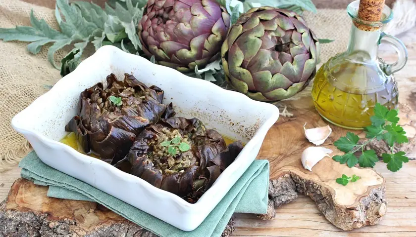 Carciofi al forno morbidi ripieni, serviti in una teglia bianca con olio d'oliva, aglio e prezzemolo.