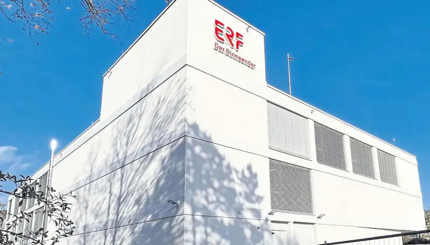 Das Gebäude von ERF, dem Sinn-Sender, unter blauem Himmel. Schatten von Bäumen tanzen auf der weißen Fassade.