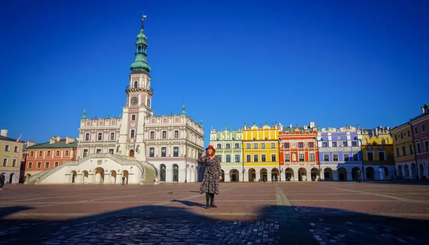 Kobieta w czerwonym kapeluszu na Rynku Wielkim w Zamościu, podziwia kolorowe kamienice i ratusz. Zamość atrakcje zachwycają.