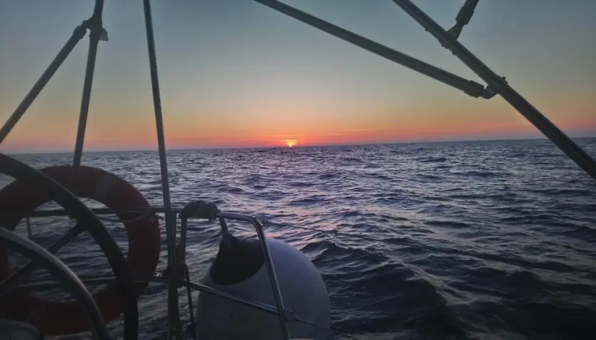 Zachód słońca nad wzburzonym oceanem atlantyckim widziany z pokładu żaglówki. Widać koło ratunkowe i ster.