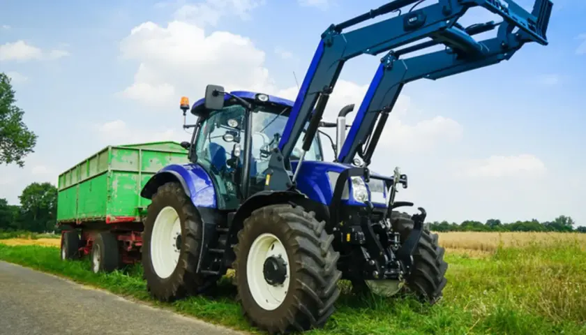 Blauer Traktor mit Frontlader und grünem Anhänger steht am Feldrand. Ein Traktor fahren ohne Landwirtschaft ist hier möglich, vielleicht für eine Baustelle.
