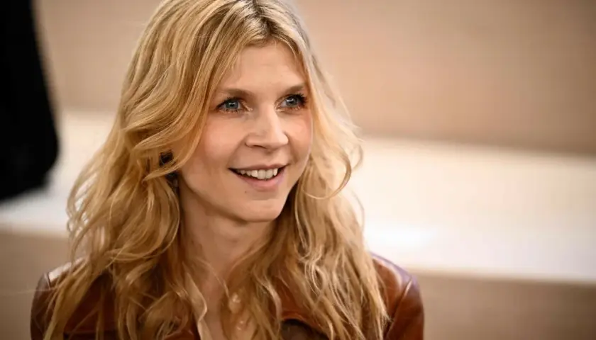 Uśmiechnięta Clémence Poésy z falującymi blond włosami i niebieskimi oczami, ubrana w brązową skórzaną kurtkę.