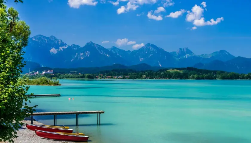 Türkisblaues Wasser am Badesee Bayern, mit roten Ruderbooten am Kiesstrand und majestätischen Alpen im Hintergrund.