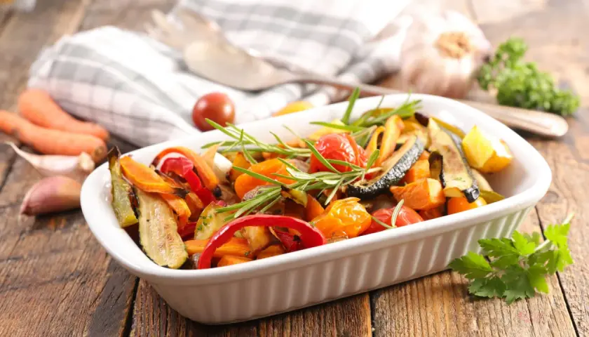 Un plato blanco lleno de coloridas verduras al horno, con romero fresco y tomates cherry.