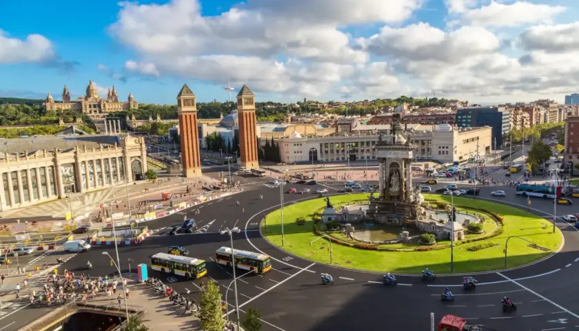 Plac Hiszpański w Barcelonie, z dwoma wieżami w tle i fontanną pośrodku. Ruch uliczny, autobusy i motocykle tworzą tętniącą życiem scenę w tym pięknym, **starym mieście Barcelona**.