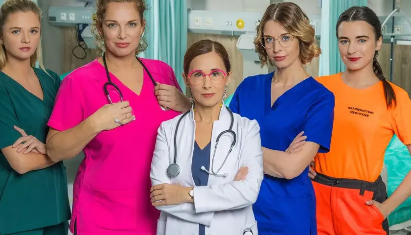 Pięć kobiet w strojach medycznych, obsada serialu "Szpital św. Anny", pozuje do zdjęcia w szpitalnej sali.