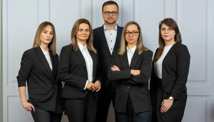 Pięć osób w garniturach, zespół prawników, stoi przed jasną ścianą.