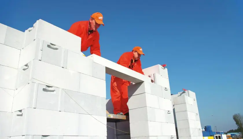 Dwóch budowlańców w pomarańczowych strojach montuje betonowy nadproże nad otworem w ścianie z białych pustaków.