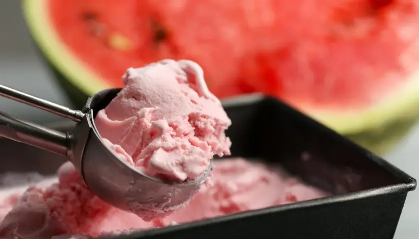 Un cucchiaio raccoglie una pallina di cremoso gelato anguria fatto in casa, con una fetta di anguria sullo sfondo.