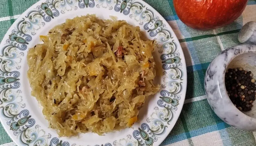Na talerzu z ozdobnym wzorem znajduje się porcja pysznej, **kapusty kiszonej gotowanej**. Obok jabłko i moździerz z pieprzem.