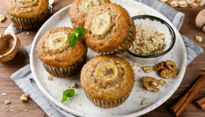 Pięć pysznych muffinek bananowych z plasterkiem banana na wierzchu, udekorowane miętą. Obok miseczka z orzechami i laska cynamonu.