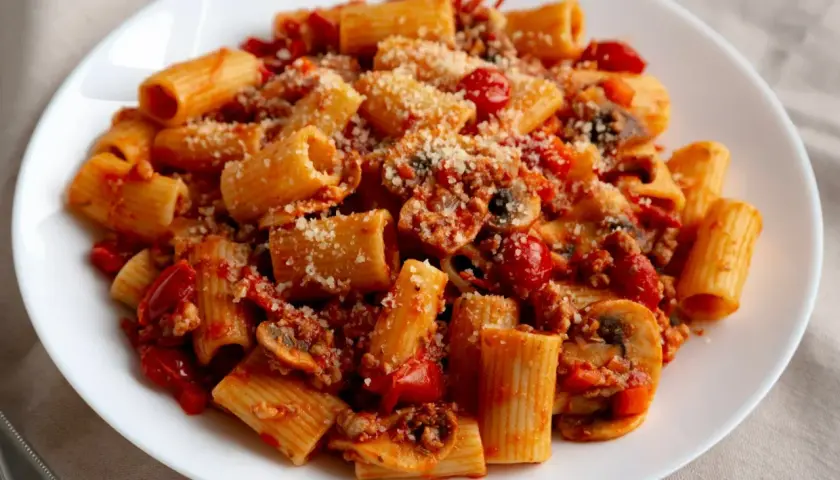 Pyszny czerwony makaron rigatoni z sosem bolońskim, pieczarkami i pomidorkami, posypany parmezanem.