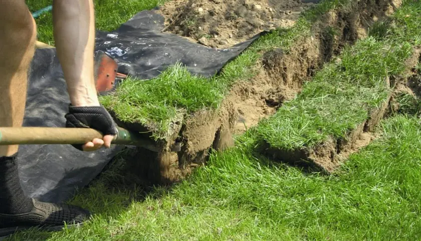 Osoba w rękawiczkach kopie łopatą rów, wycinając darnie. Prace ogrodowe w pełnym słońcu.