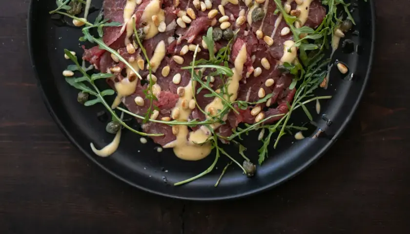 Rinder Carpaccio mit Pinienkernen, Kapern und Rucola auf schwarzer Platte, beträufelt mit einer cremigen Sauce.