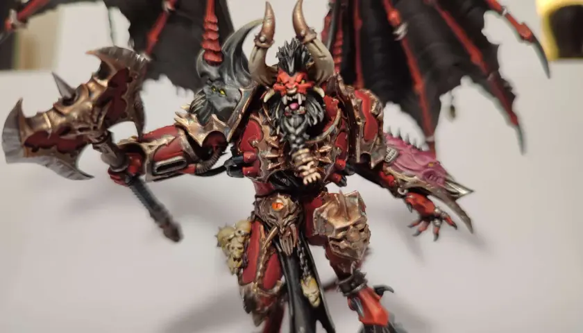 Groźny chaos daemon prince z czerwono-złotą zbroją, czarnymi skrzydłami i toporem.