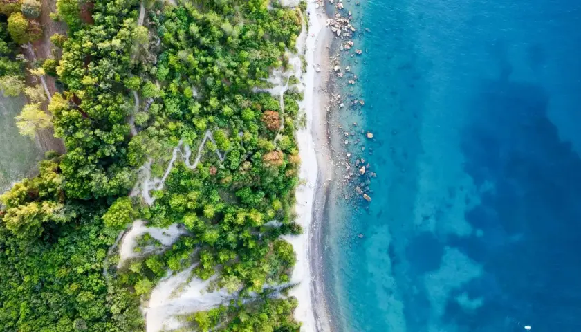 Luftaufnahme eines Küstenstreifens mit üppigem Grün, einem Kiesstrand und dem türkisfarbenen Ljubljana Meer.