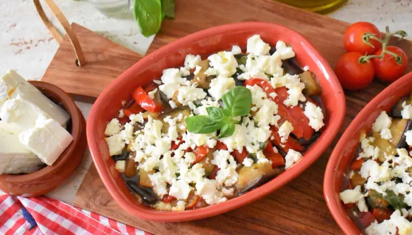 Pyszna pieczona feta z warzywami, podana w glinianym naczyniu, z dodatkiem świeżej bazylii i pomidorków.