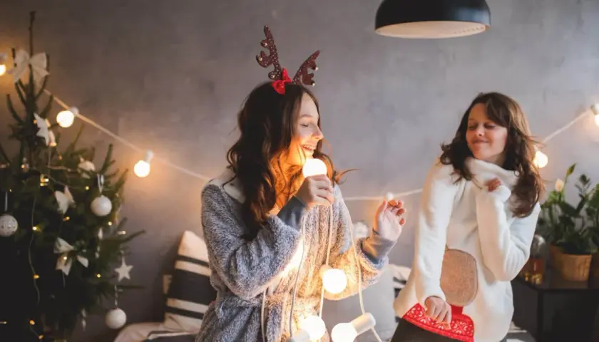 Due ragazze cantano canzoni di Natale in inglese con testo, tenendo una lampadina come microfono, circondate da luci natalizie e un albero addobbato.