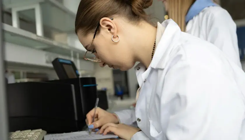 Studentka w białym fartuchu laboratoryjnym pisze notatki w zeszycie, obok klawiatury i telefonu.