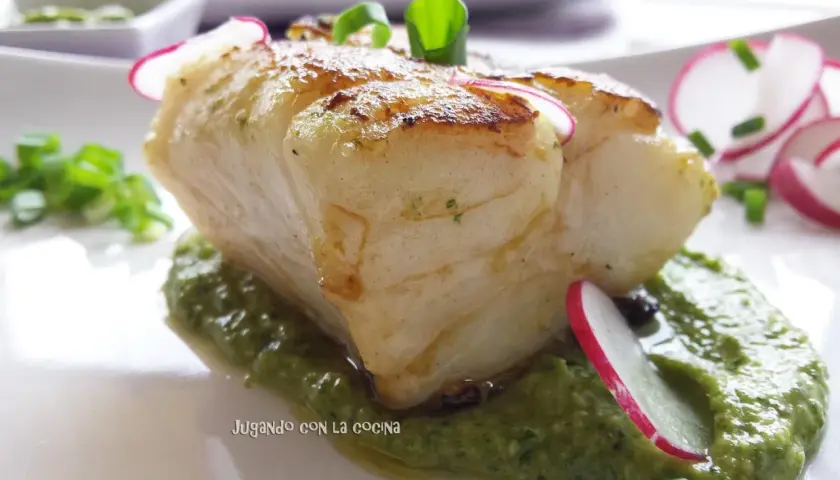 Un trozo de pescado blanco y tierno reposa sobre una cama de vibrante mojo verde, adornado con rodajas de rábano y cebolleta.