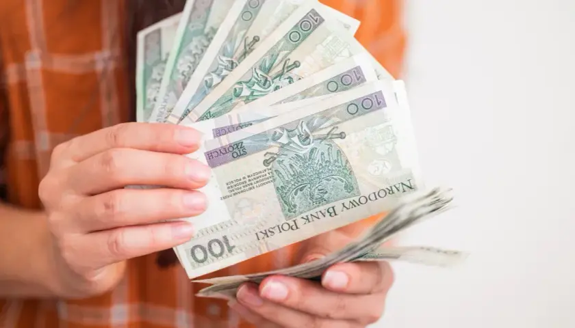 Dłonie trzymają wachlarz banknotów 100 zł. Taka kwota to już całkiem spora kuroniówka na drobne wydatki.