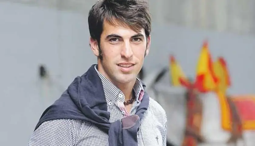 Roberto Armendáriz, con su camisa a cuadros y jersey sobre los hombros, sonríe. Al fondo, un caballo engalanado con banderas.