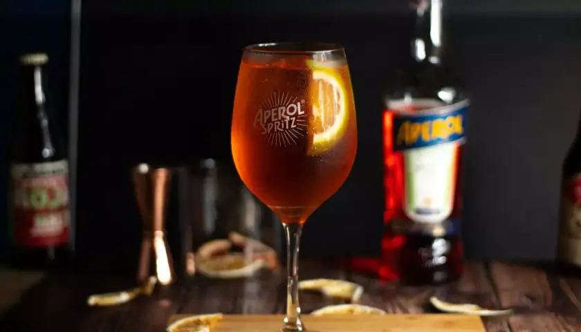 Aperol Spritz w kieliszku z plasterkiem pomarańczy, obok butelka Aperol i inne dodatki.