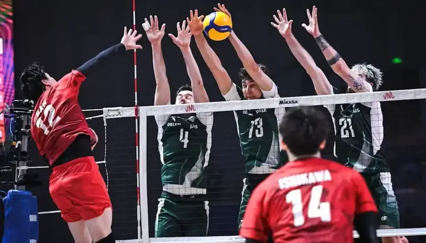 Zacięta akcja siatkarska: polska Japonia w akcji. Zawodnik w czerwonej koszulce atakuje, blokujący w zielonych strojach próbują zatrzymać piłkę.