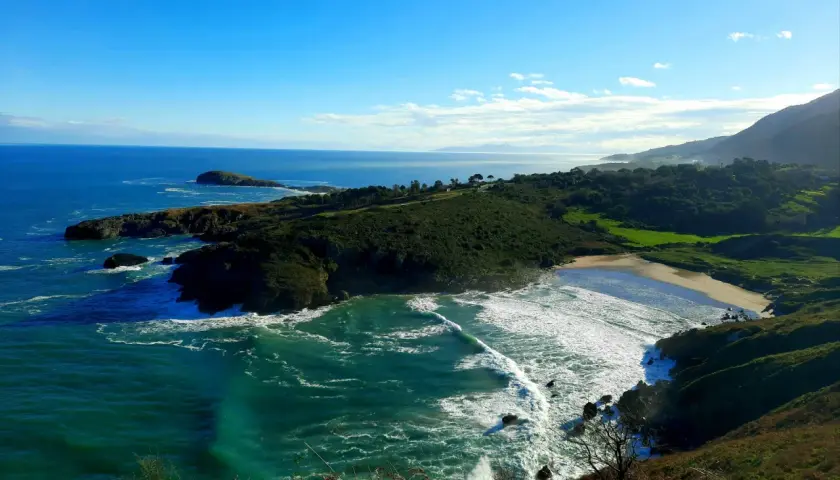Vista panorámica de la playa de Sonabia, con olas rompiendo en la costa rocosa y un mar azul intenso bajo un cielo despejado.