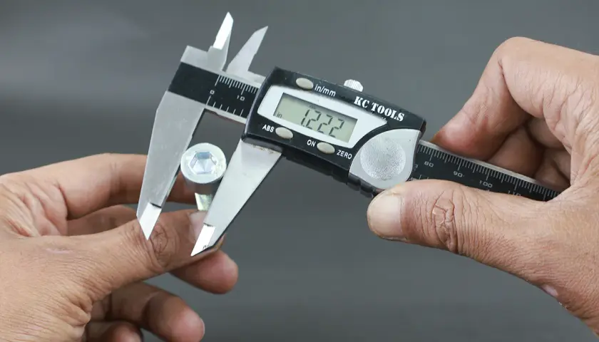 Manos sujetan un tornillo hexagonal y un calibre digital para su calibre medicion. La pantalla muestra 1.222.