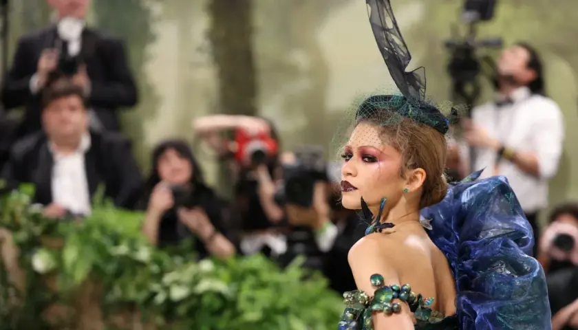 Una modelo con un elaborado atuendo de **Met Gala theme**, con velo y maquillaje dramático, posa rodeada de fotógrafos y vegetación.