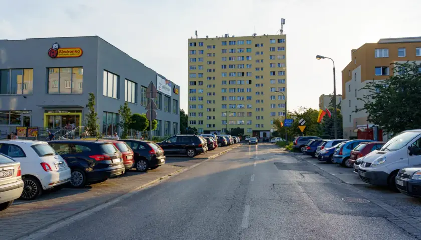 Uliczka z zaparkowanymi samochodami obok sklepu Biedronka, gdzie logo przypomina uśmiechniętego ursusa niedźwiadka. W tle wysoki, żółty blok mieszkalny.