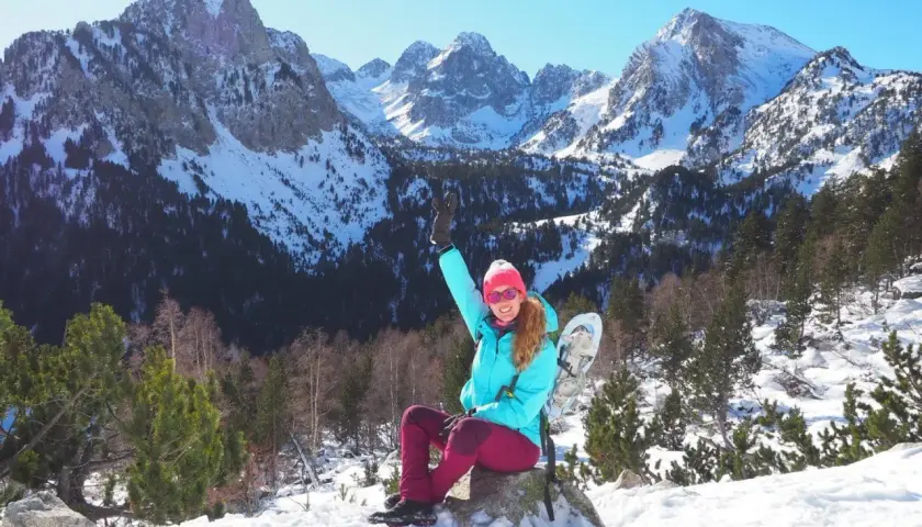 Mujer con raquetas de nieve saluda en un paisaje nevado de **Pallars Sobirà**. Montañas imponentes y bosques cubiertos de nieve crean un telón de fondo espectacular.