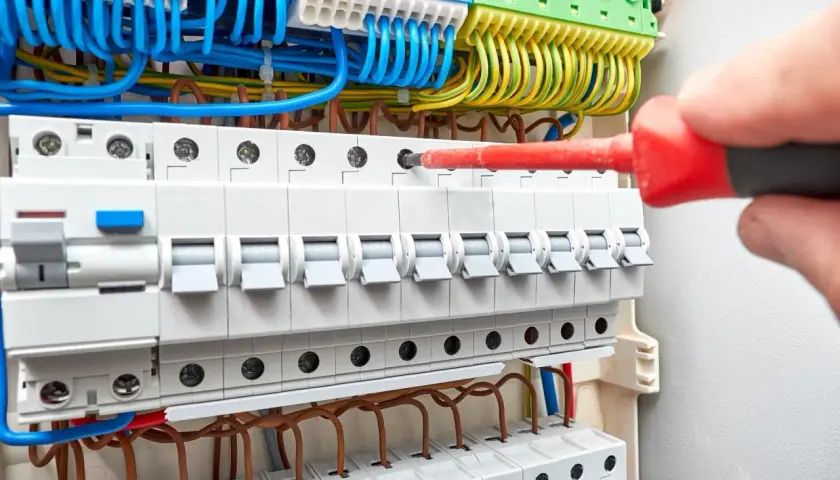 Ręka z czerwonym śrubokrętem przykręca przewody w skrzynce elektrycznej. Trwa wymiana instalacji elektrycznej.