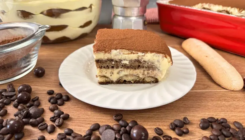 Una fetta di tiramisù con uova pastorizzate, cosparso di cacao, su un piatto bianco, con chicchi di caffè e cioccolatini sparsi.