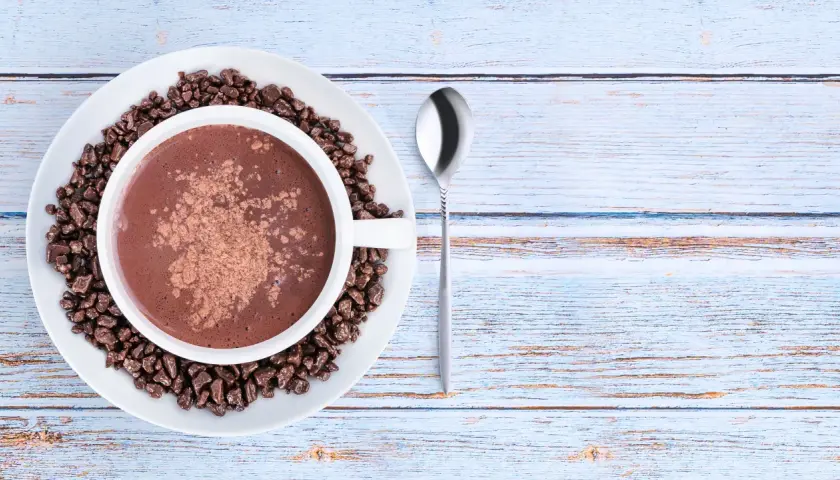 Tazza di cioccolata calda con cacao amaro in polvere e chicchi di cioccolato, su un piatto bianco, accanto a un cucchiaino. Le proprietà del cacao amaro sono note per il benessere.