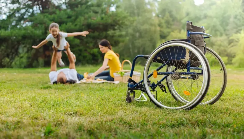 Rodzina na pikniku, ojciec podrzuca córkę, matka przygotowuje jedzenie. Obok wózek inwalidzki, symbol wsparcia i świadczenie pielęgnacyjne 2026.