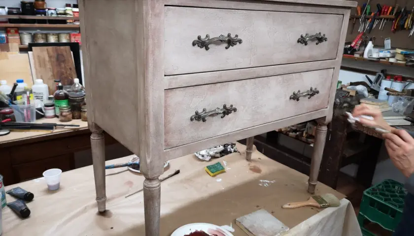 Un mobile antico in fase di restauro, con cassetti e pomelli decorati. Attrezzi e materiali da lavoro circondano il pezzo.